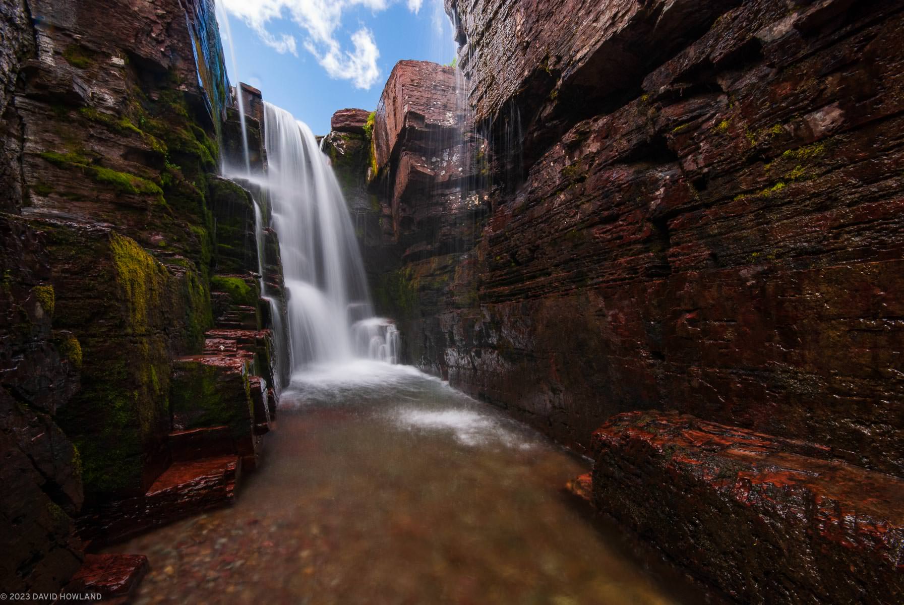 A photo of a narrow waterfall cascading over a rocky ledge into a narrow slot canyon in Glacier National Park