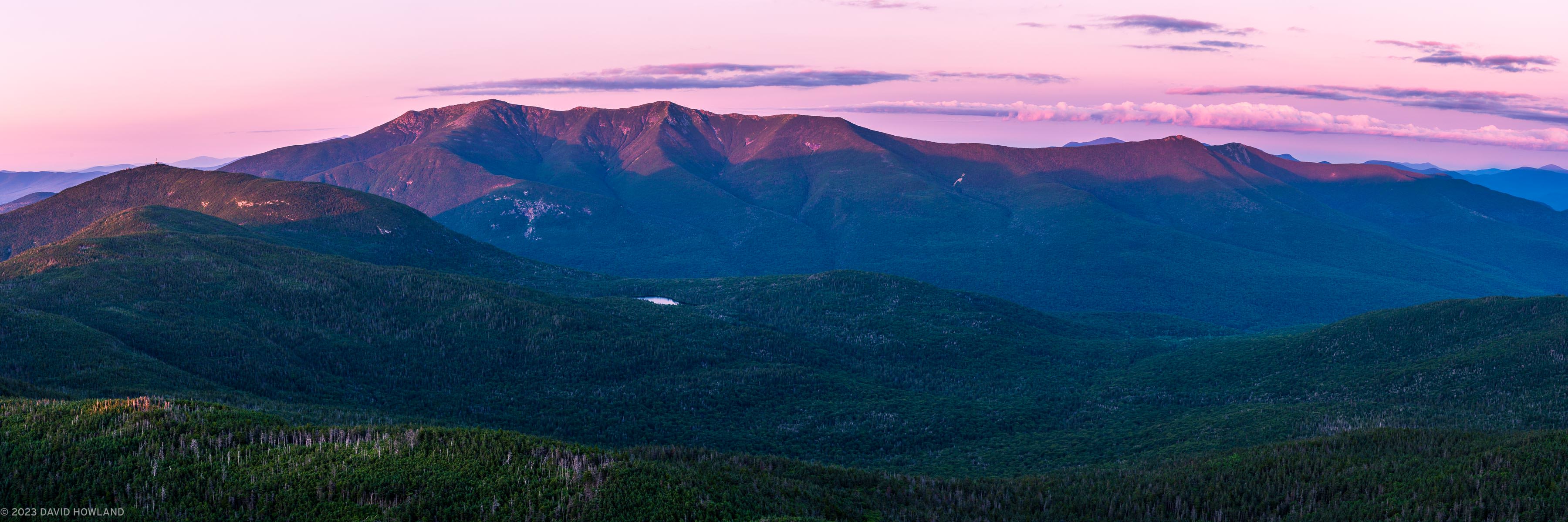 The image shows a panoramic view of mountain ranges at sunset, captured in New Hampshire's Franconia Notch region. The foreground features forested slopes covered in dense evergreen trees, while the dramatic peaks of the mountains rise against a pink and purple twilight sky. A small lake or pond is visible nestled in the valley between the mountains. The scene is captured during summer, with lush vegetation throughout. The composition emphasizes the scale and grandeur of the landscape through its wide format and layered perspective, from the foreground forest through multiple mountain ridges receding into the distance. The pink-hued sky creates a striking contrast with the deep blue-green tones of the mountains and forest below.