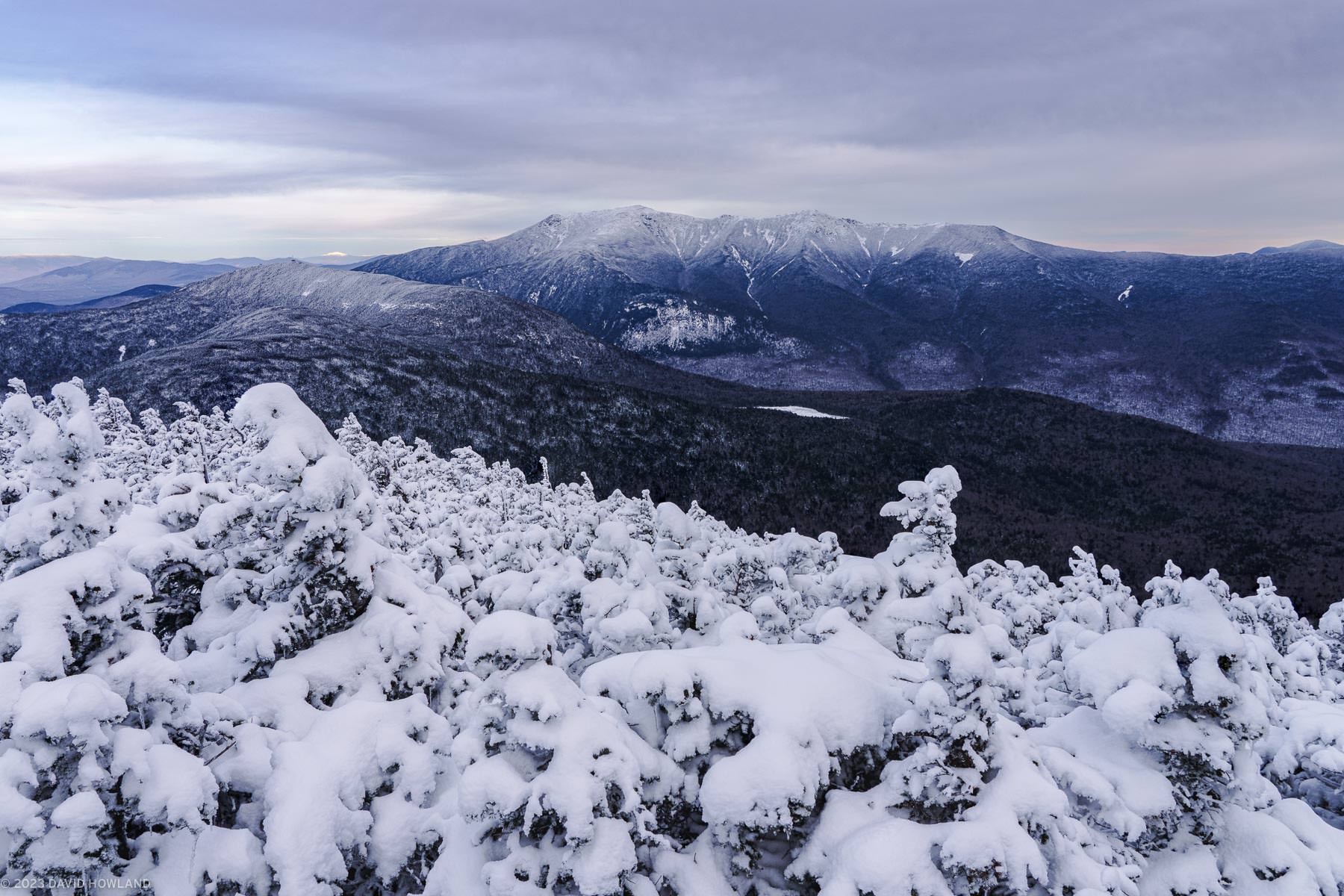 A photo of snowy trees in front of the snow-covered peaks of Franconia Notch in the White Mountains of New Hampshire