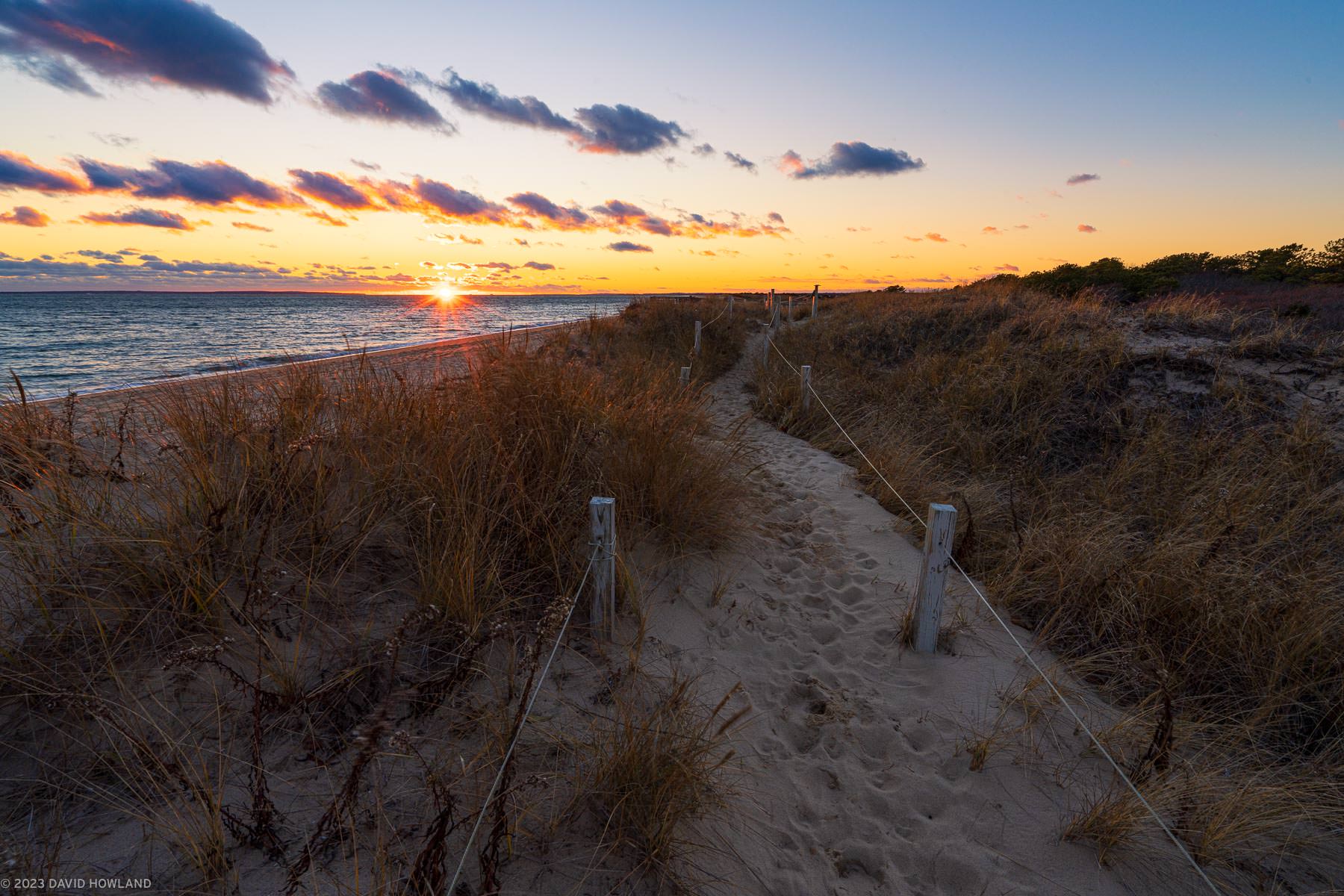 A photo of the sun setting behind the Atlantic Ocean and a sandy path through the dune grass of South Cape Beach State Park on Cape Cod.