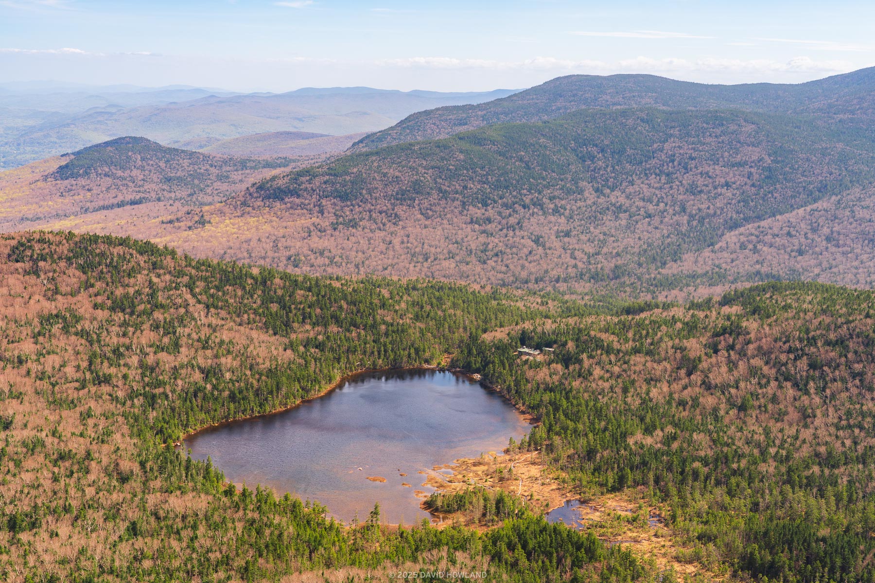 This landscape photograph captures a pristine mountain lake nestled within New England's rolling hills during spring, with the forested landscape displaying a mix of emerging green foliage and bare deciduous trees creating a patchwork of colors across the undulating terrain. The circular lake sits like a dark mirror in the valley below, surrounded by multiple ridgelines that fade into distant blue-gray mountain silhouettes under soft, diffused lighting.