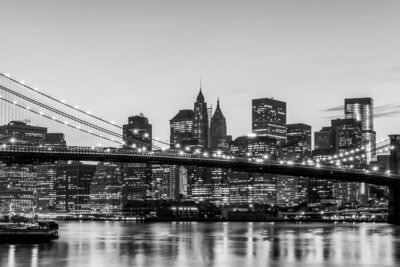 A black and white panorama photo of the Brooklyn Bridge in front of the illuminated skyscrapers of downtown New York City at night.