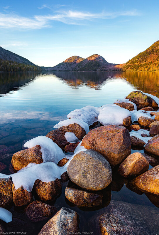 Calm pond with ice-capped rocks in the foreground and two rounded mountains reflected in the still water at sunset.