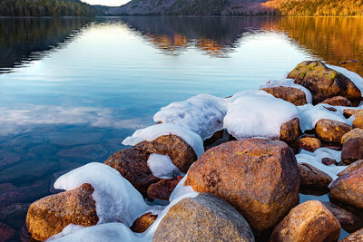 Calm pond with ice-capped rocks in the foreground and two rounded mountains reflected in the still water at sunset.
