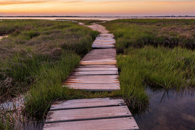 A photo of a boardwalk in a salt marsh under glowing orange skies at sunset in Barnstable, Massachusetts on Cape Cod.